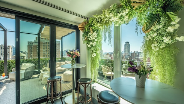Un balcon moderne avec une porte vitrée, une table ronde, deux tabourets de bar et un vase de fleurs.