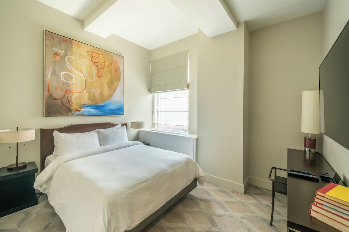 Habitación de hotel con una cama grande, un escritorio con lámpara, una ventana con persianas y un cuadro en la pared.