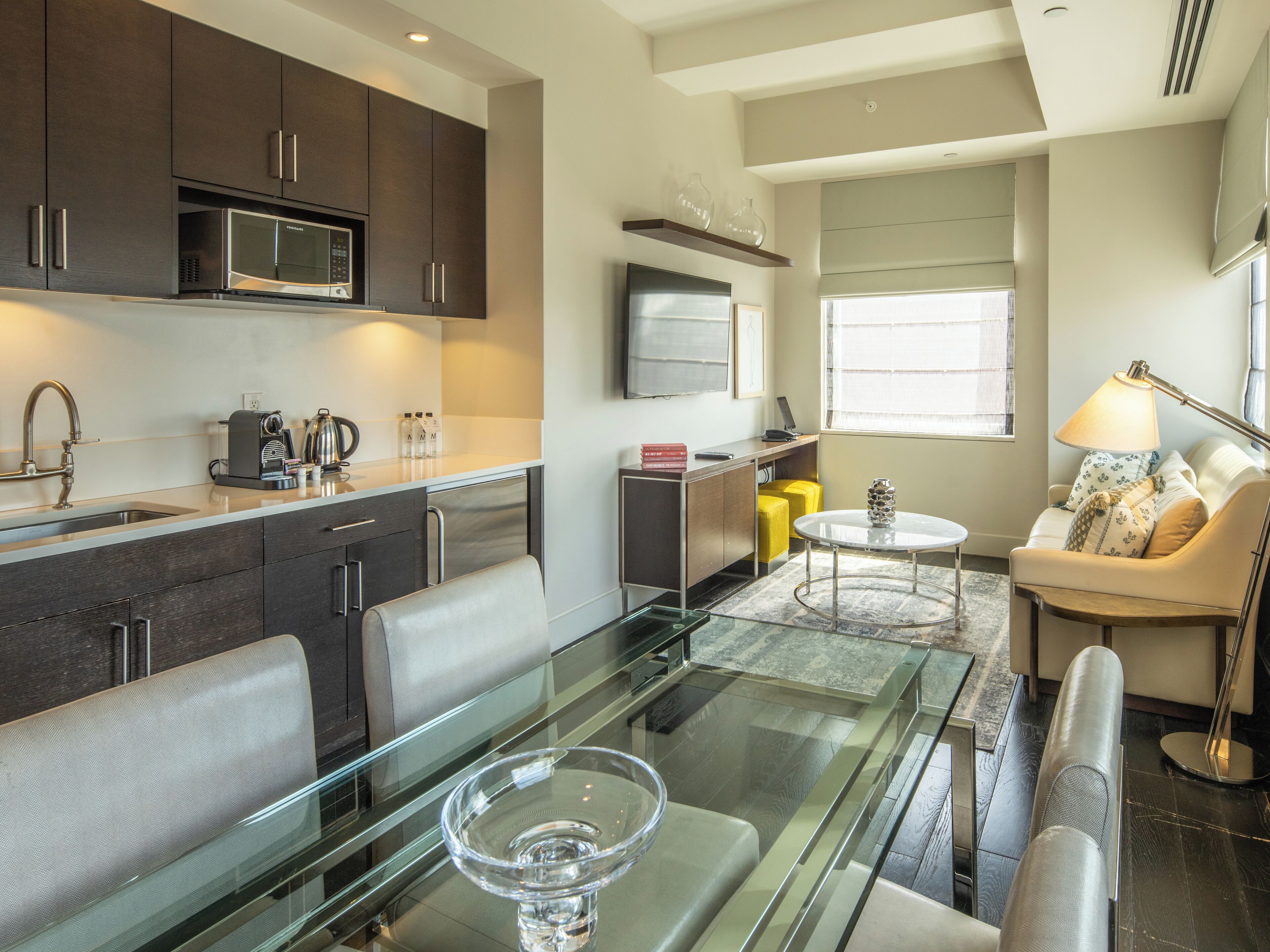 A modern kitchen with dark cabinets, a microwave, and a coffee maker. There is a dining area with a glass table and chairs, and a living area with a sofa and a lamp.