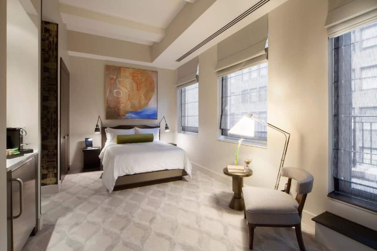 Habitación de hotel con cama, mesita de noche, lámpara, silla y una obra de arte en la pared.