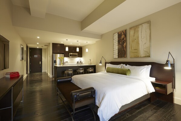 A modern hotel room with a large bed, a desk, a bench, a kitchenette, and artwork on the walls.
