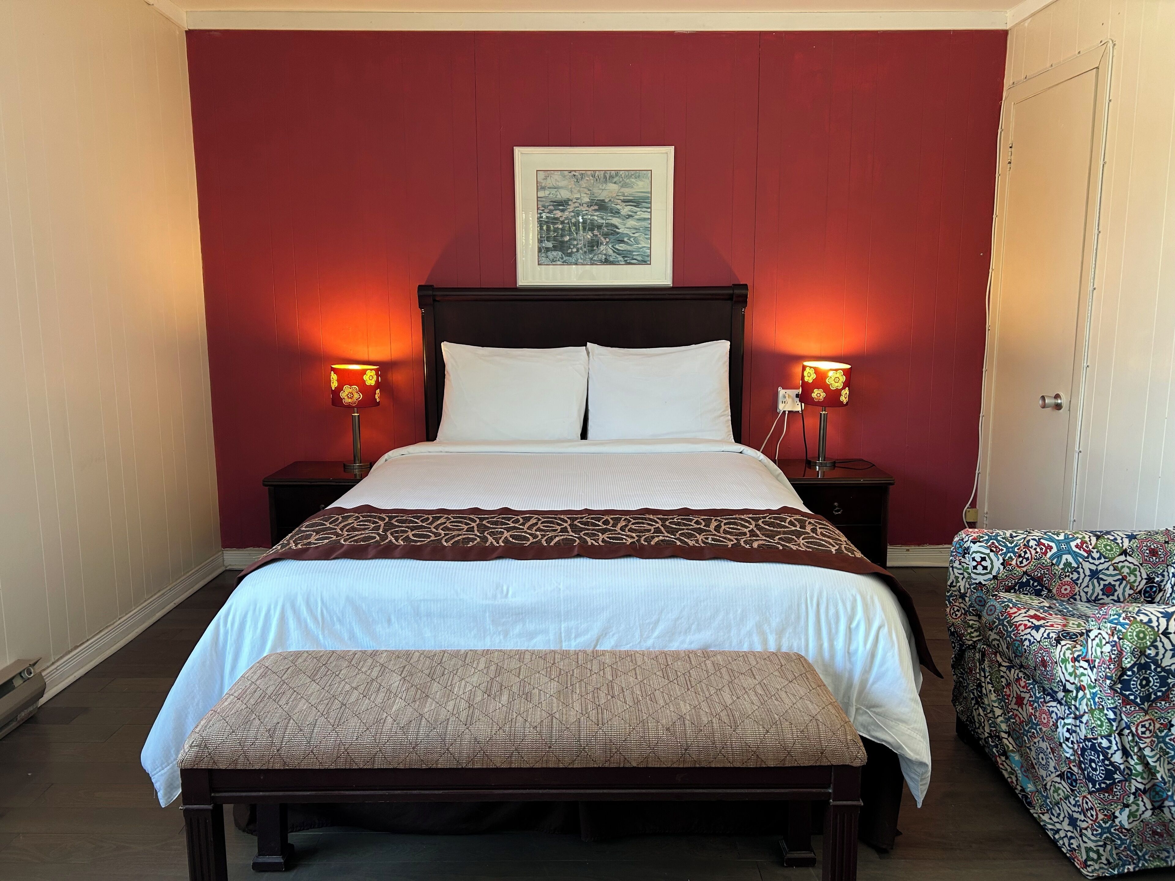 A neatly made bed with a patterned bedspread, two bedside lamps, a framed picture above the bed, and a bench with a cushion in front of the bed.