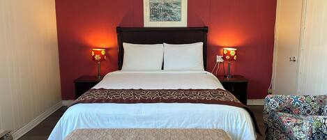 A neatly made bed with a patterned bedspread, two bedside lamps, a framed picture above the bed, and a bench with a cushion in front of the bed.