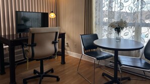 A hotel room with a TV, two chairs, a small table with a vase of flowers, and a window with patterned curtains.