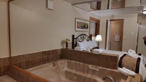 A hotel bathroom with a large bathtub, a bed with white linens, a bedside table with a lamp, and a mirror above the bathtub.