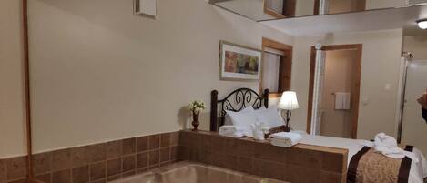 Un baño de hotel con una bañera grande, una cama con sábanas blancas, una mesita de noche con lámpara y un espejo sobre la bañera.