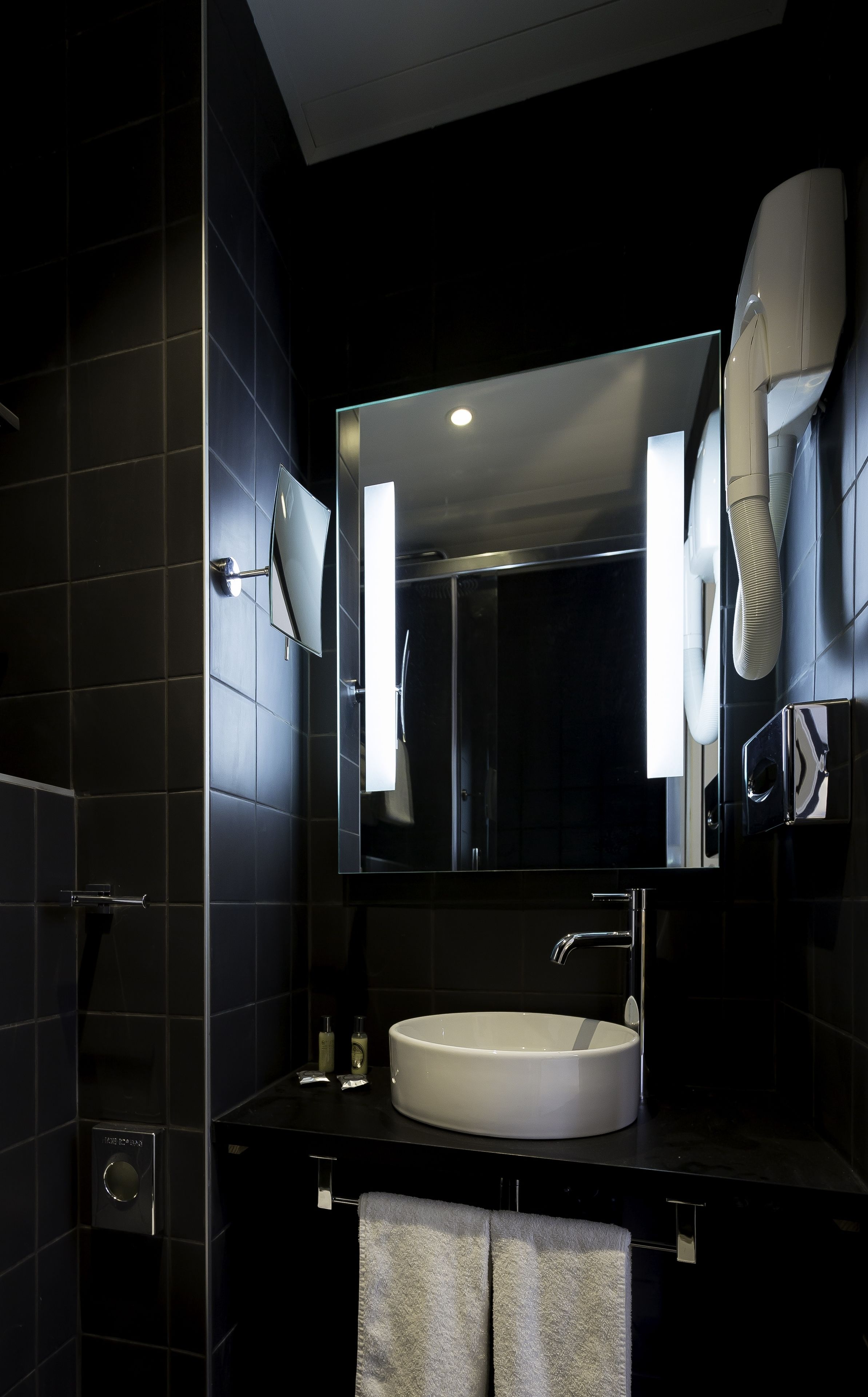 Ein modernes Badezimmer mit einem großen Spiegel, einem weißen Waschbecken und einem an der Wand befestigten Föhn.