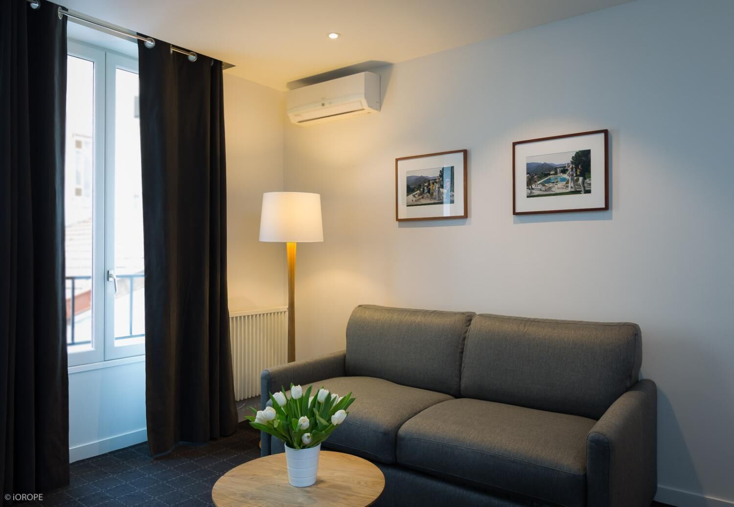 Een moderne woonkamer met een grijze bank, een ronde houten salontafel met een witte vaas met bloemen, een vloerlamp en twee ingelijste foto's aan de muur.
