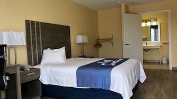 A hotel room with a bed, bedside tables, a desk, a lamp, a mirror, and a bathroom visible through an open door.