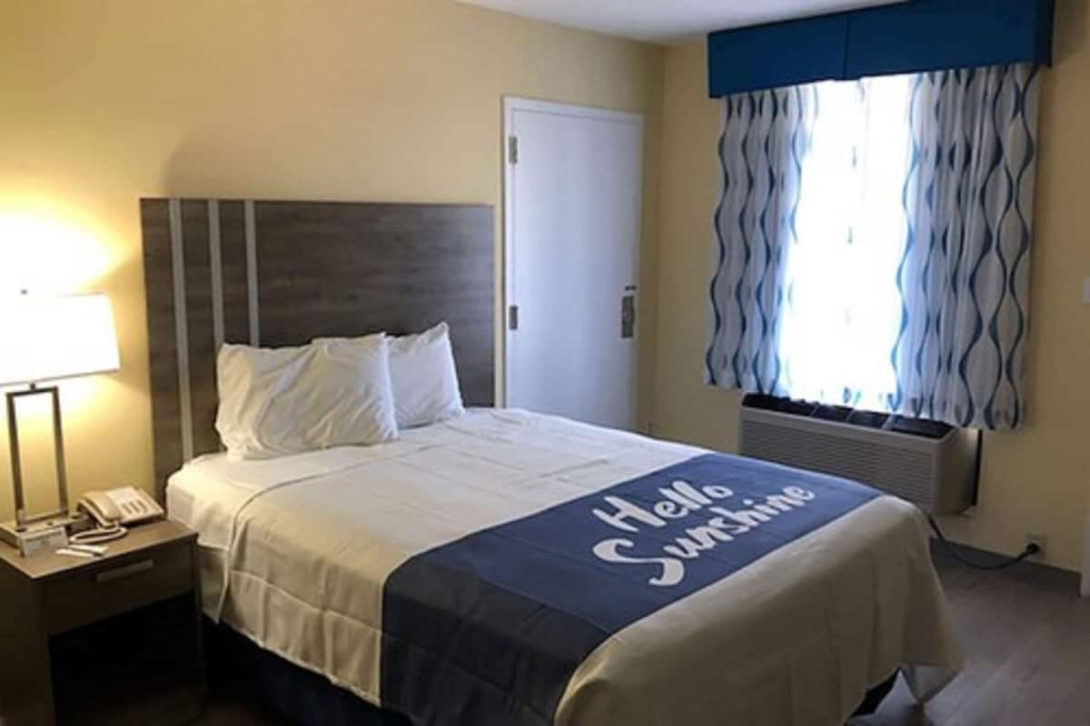 A hotel room with a bed, a bedside lamp, a telephone, a window with curtains, and a wall-mounted air conditioner.