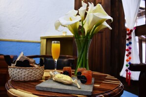 A dining table with a woven basket, a glass of orange juice, a vase with white flowers, a carved wooden spoon, and two jars of jam.