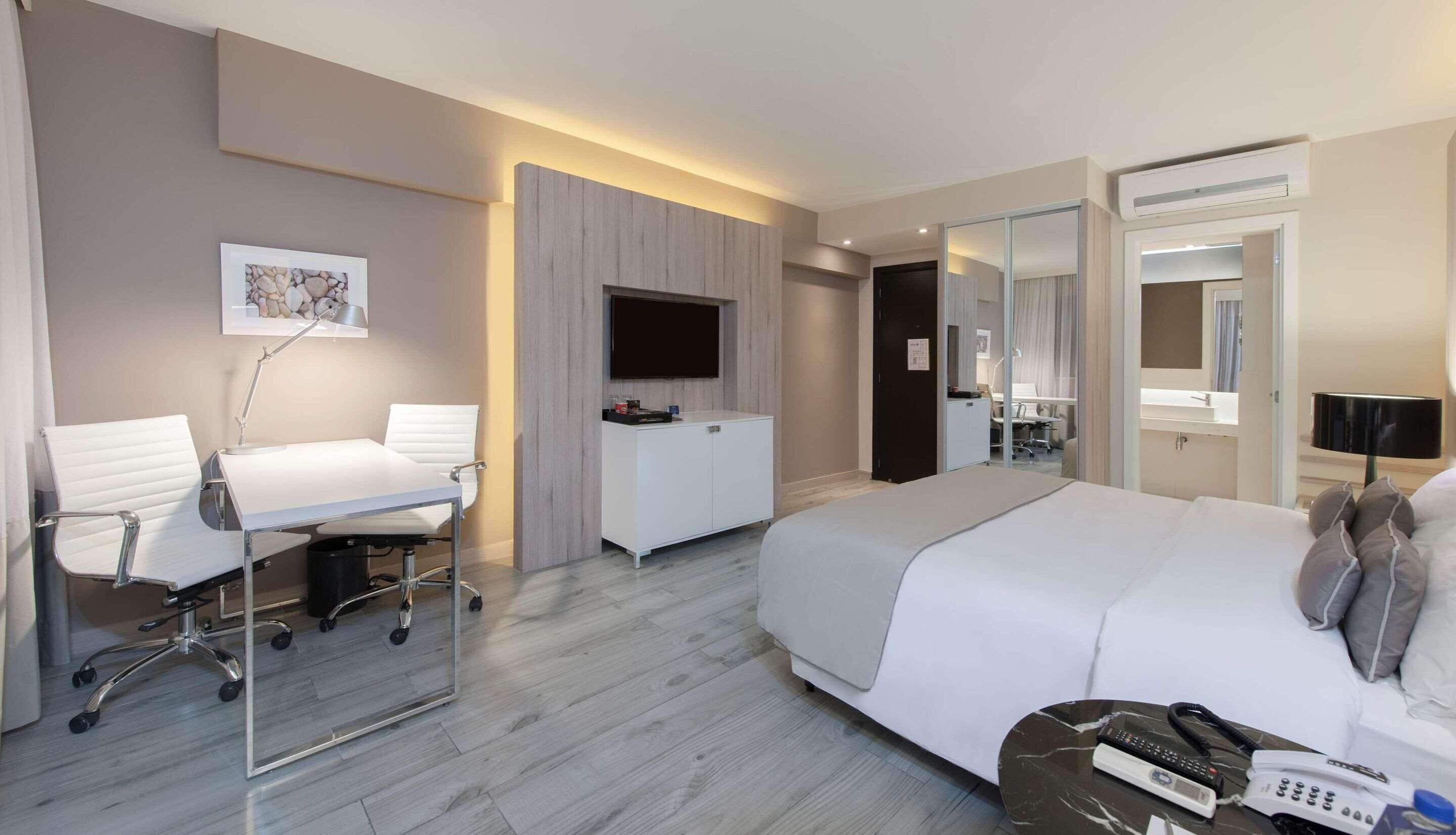 A modern hotel room with a bed, a desk with a chair, a TV, a mirror, and a bathroom visible through a glass door.