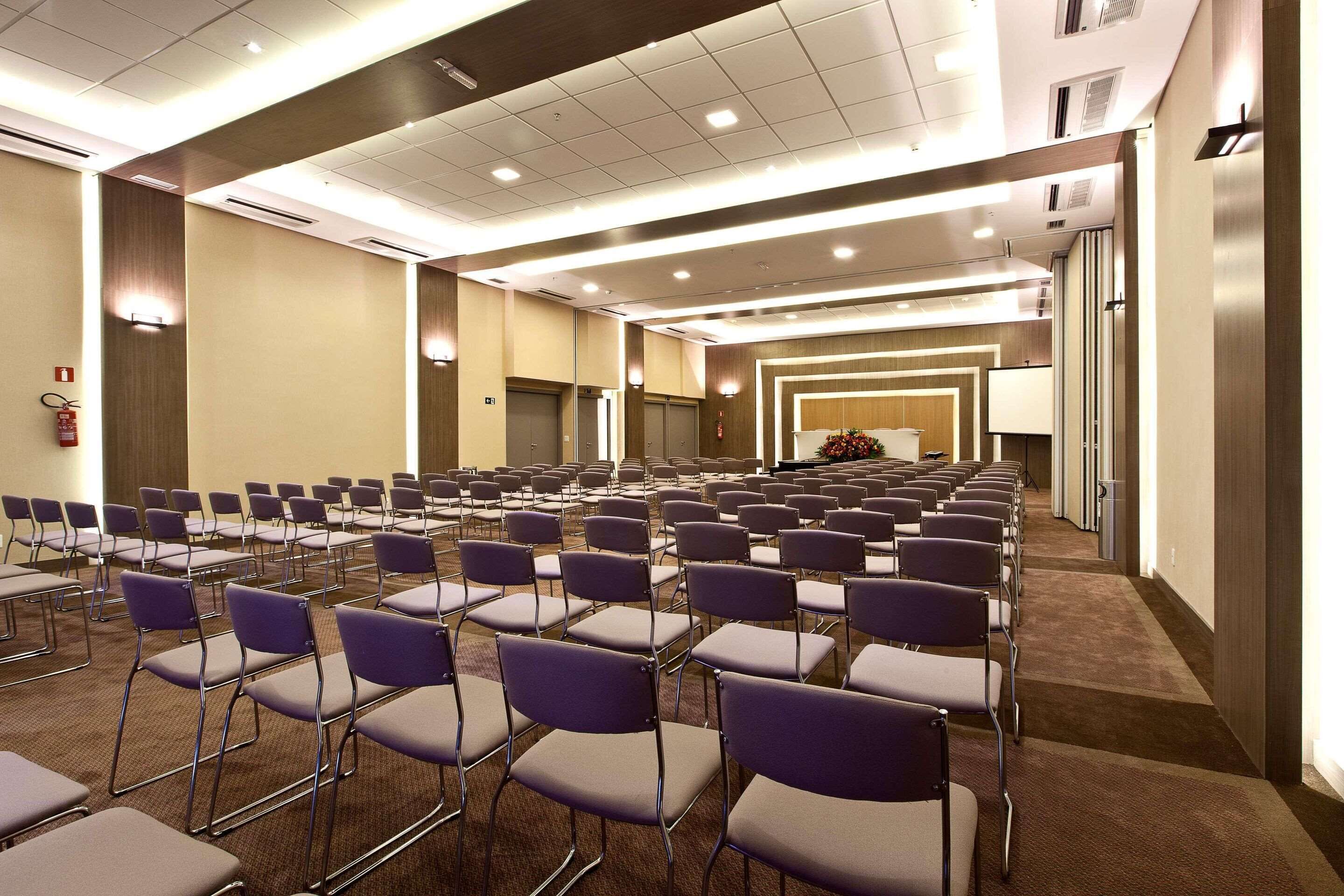 Un salón de conferencias con filas de sillas y mesas, equipado con luces en el techo y un extintor.