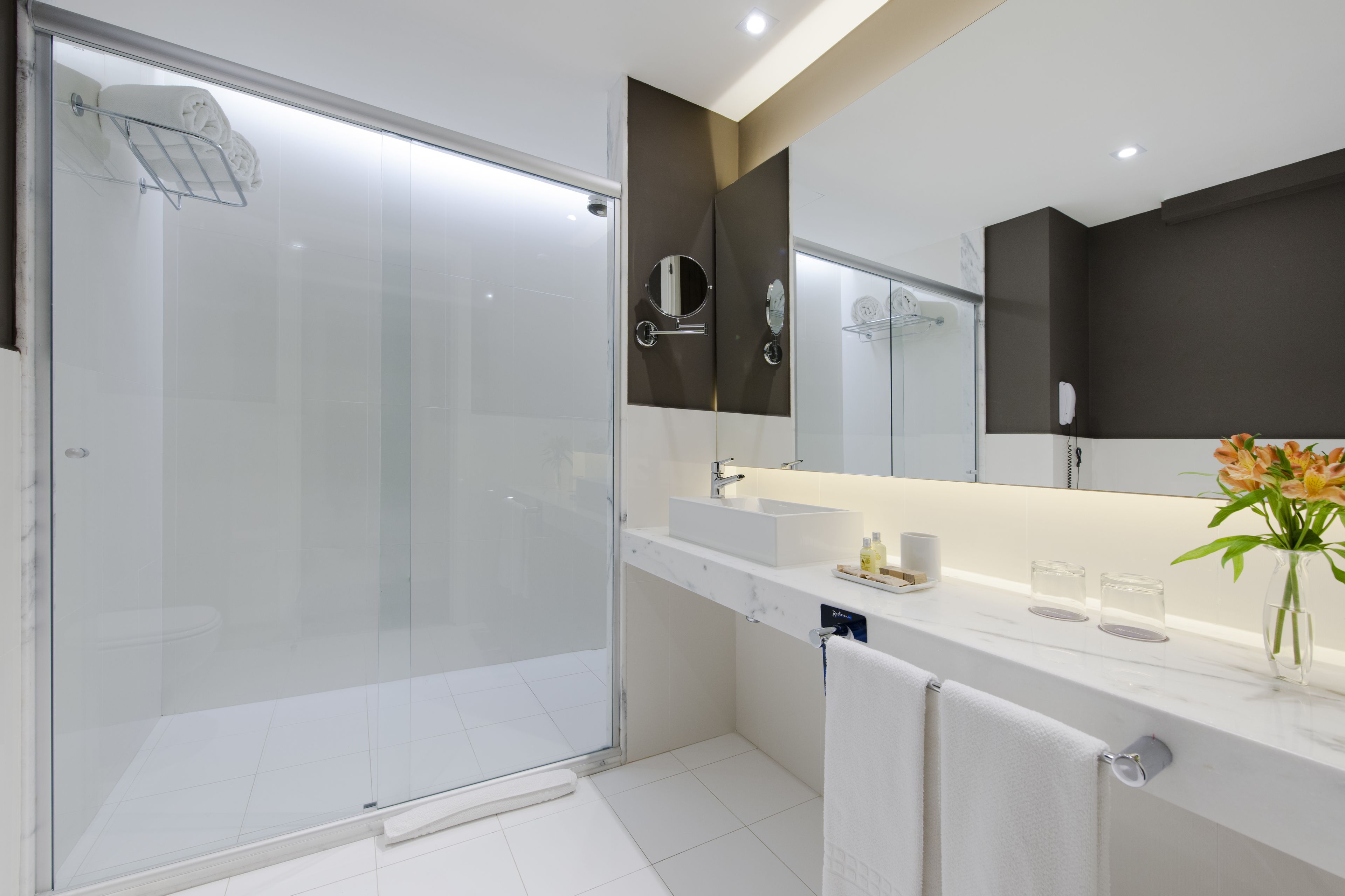 A modern bathroom with a glass-enclosed shower, a large mirror, and a white vanity with a vase of flowers.