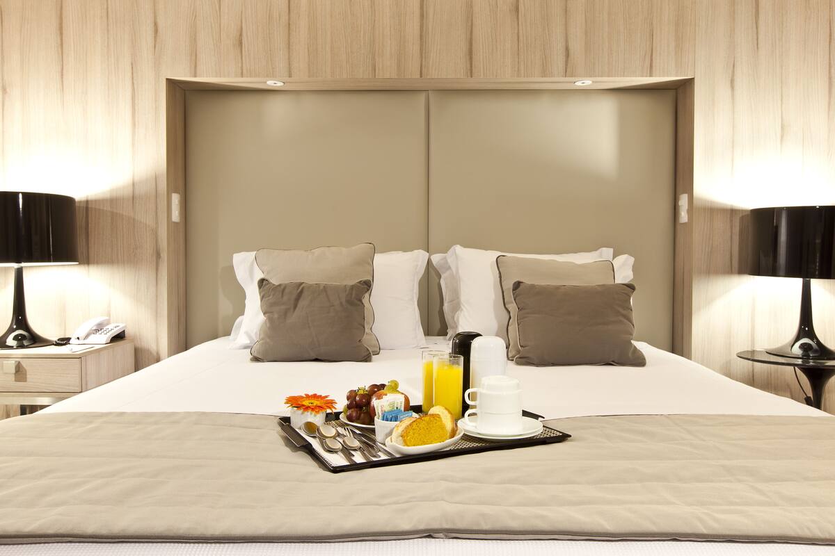A neatly made bed with a tray of breakfast items, including toast, fruit, and juice, in a modern hotel room.