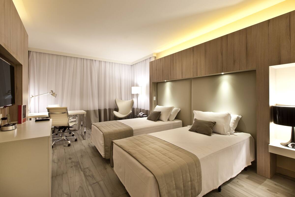 A hotel room with two beds, a desk, a chair, a TV, and a lamp.
