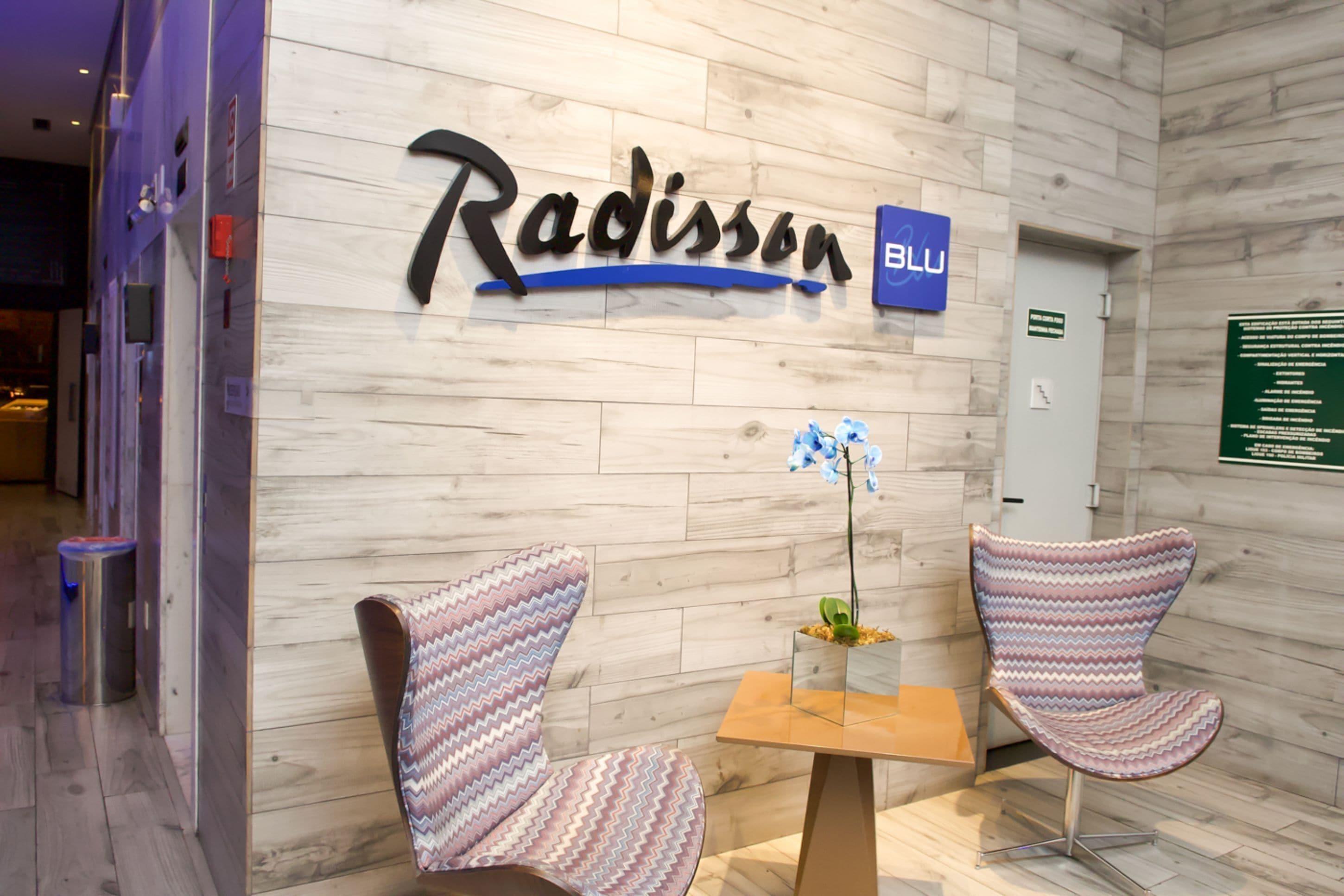 Uma área de recepção com parede de madeira, uma placa com a inscrição "Radisson BLU", duas cadeiras estampadas e uma pequena mesa com uma flor.