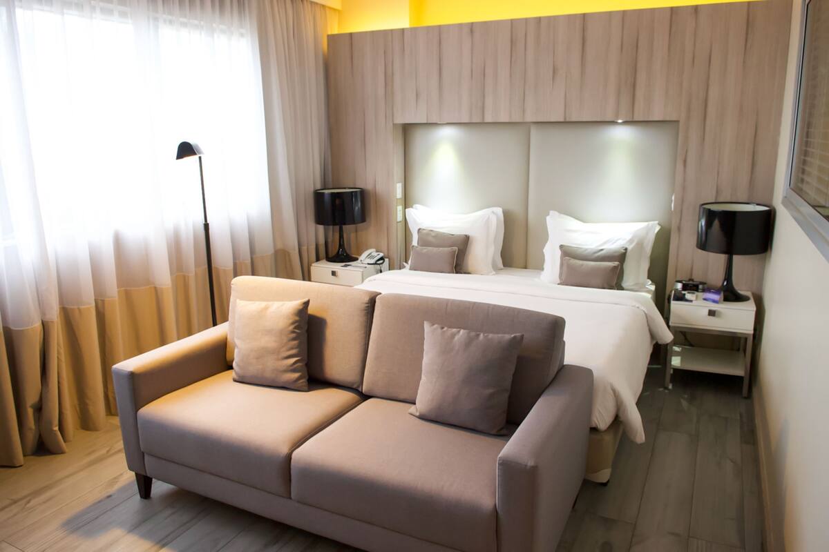 A hotel room with a bed, a sofa, two bedside tables, lamps, and a window with curtains.