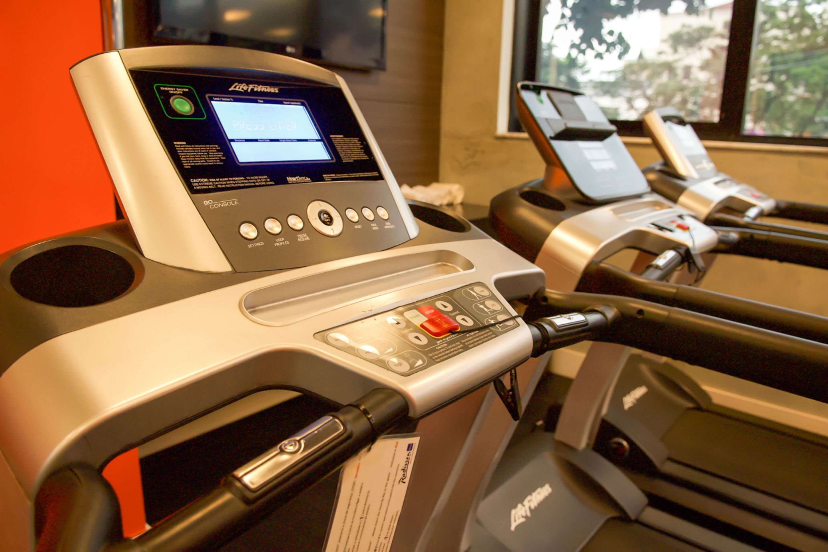 A treadmill with a digital display and control panel, located in a room with a window and exercise equipment.