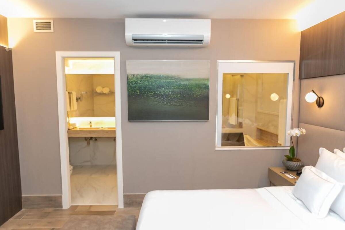 A modern hotel room with a bed, a bathroom with a shower, a mounted air conditioner, and a framed landscape painting.
