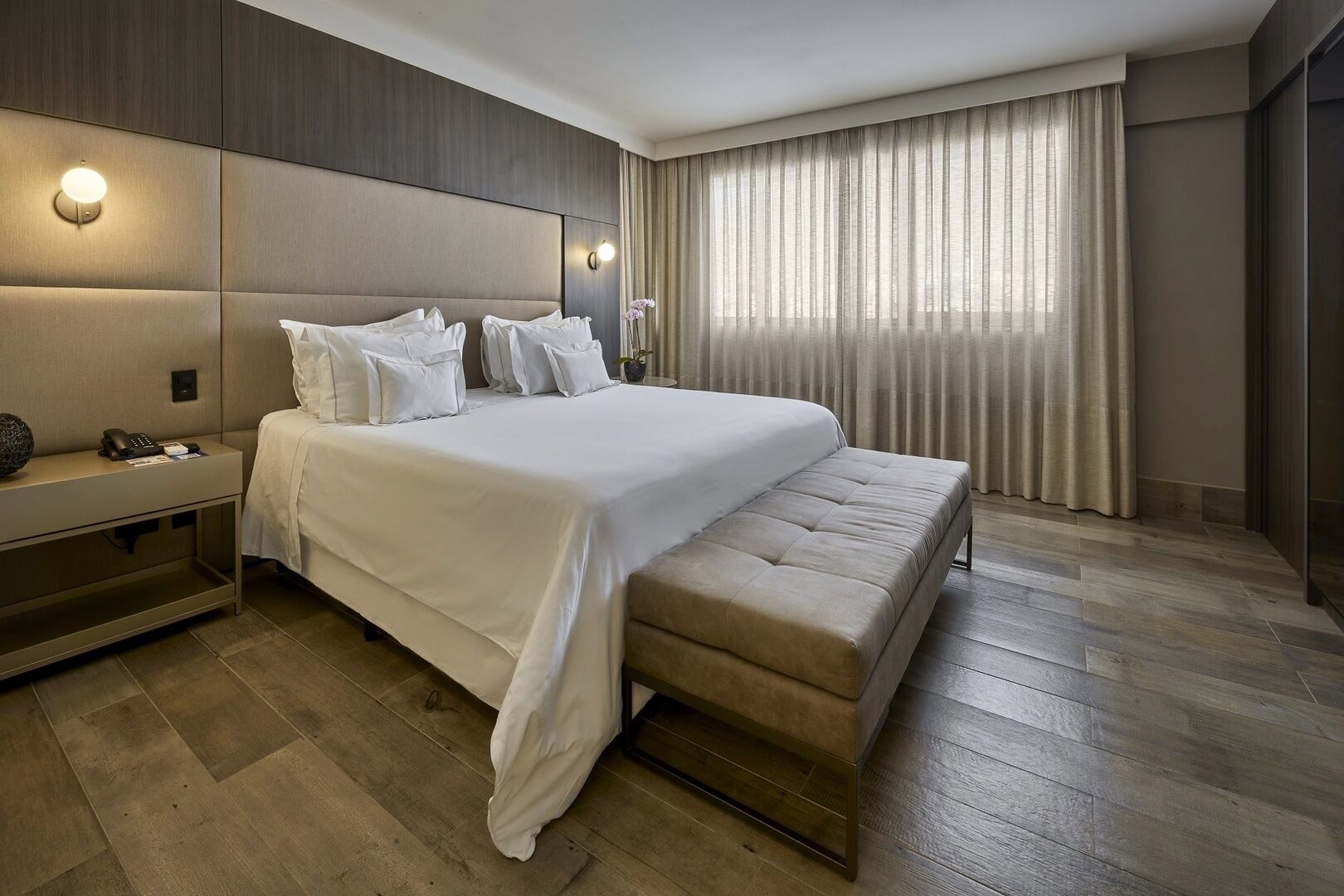 A modern hotel room with a large bed, a bedside table with a lamp, and a large window with curtains.
