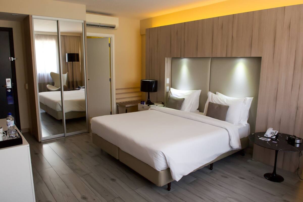 A hotel room with a large bed, a mirror, a bedside table with a lamp, and a small table with a phone and remote control.