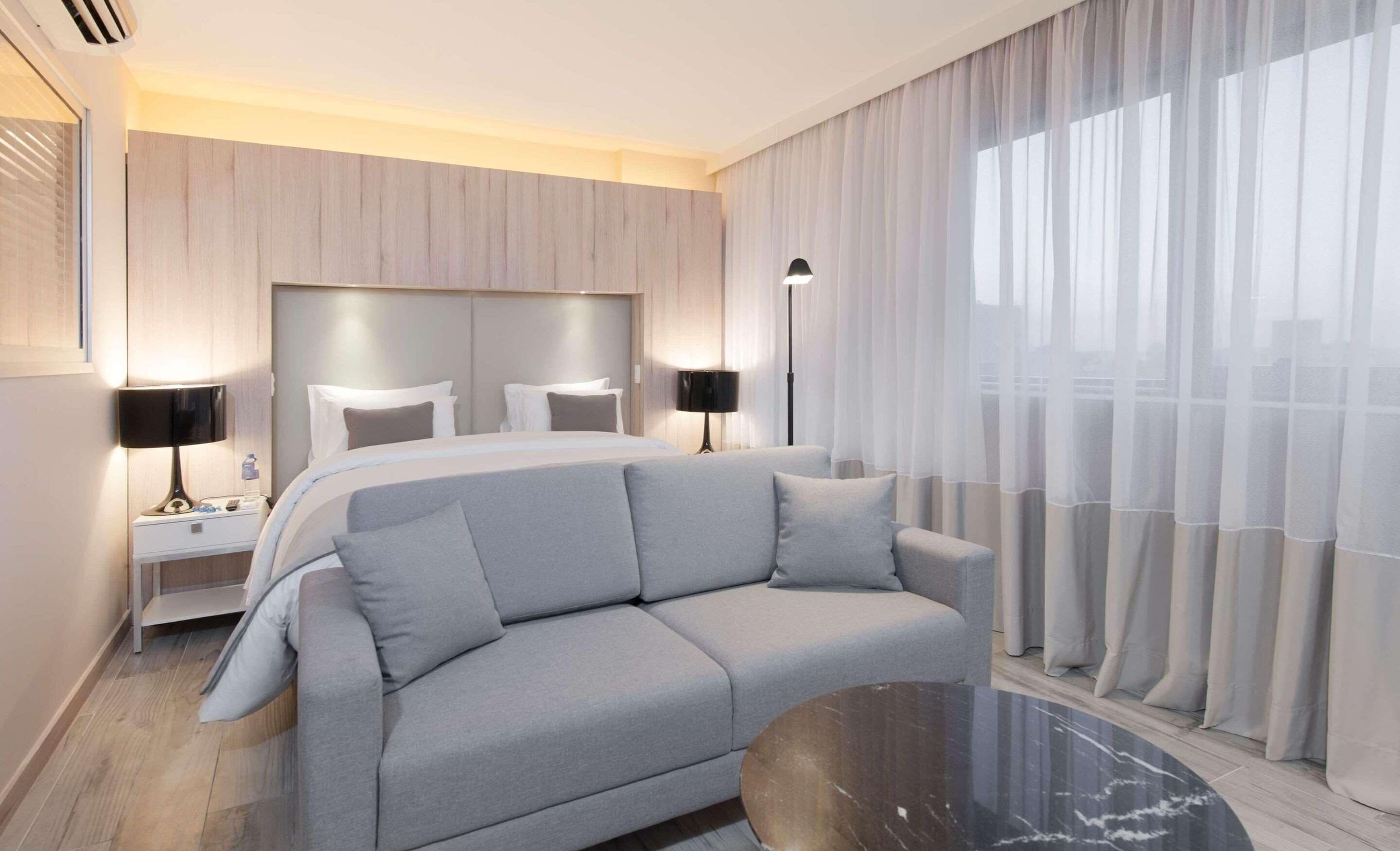Una habitación de hotel moderna con una cama grande, un sofá gris, una mesita de noche con lámpara y cortinas traslúcidas.