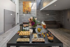 Una mesa de buffet con diversos alimentos y un centro de flores en un interior moderno con suelo de madera y un cuadro en la pared.