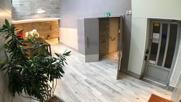 A modern lobby with a wooden floor, a reception desk, a potted plant, and an elevator.