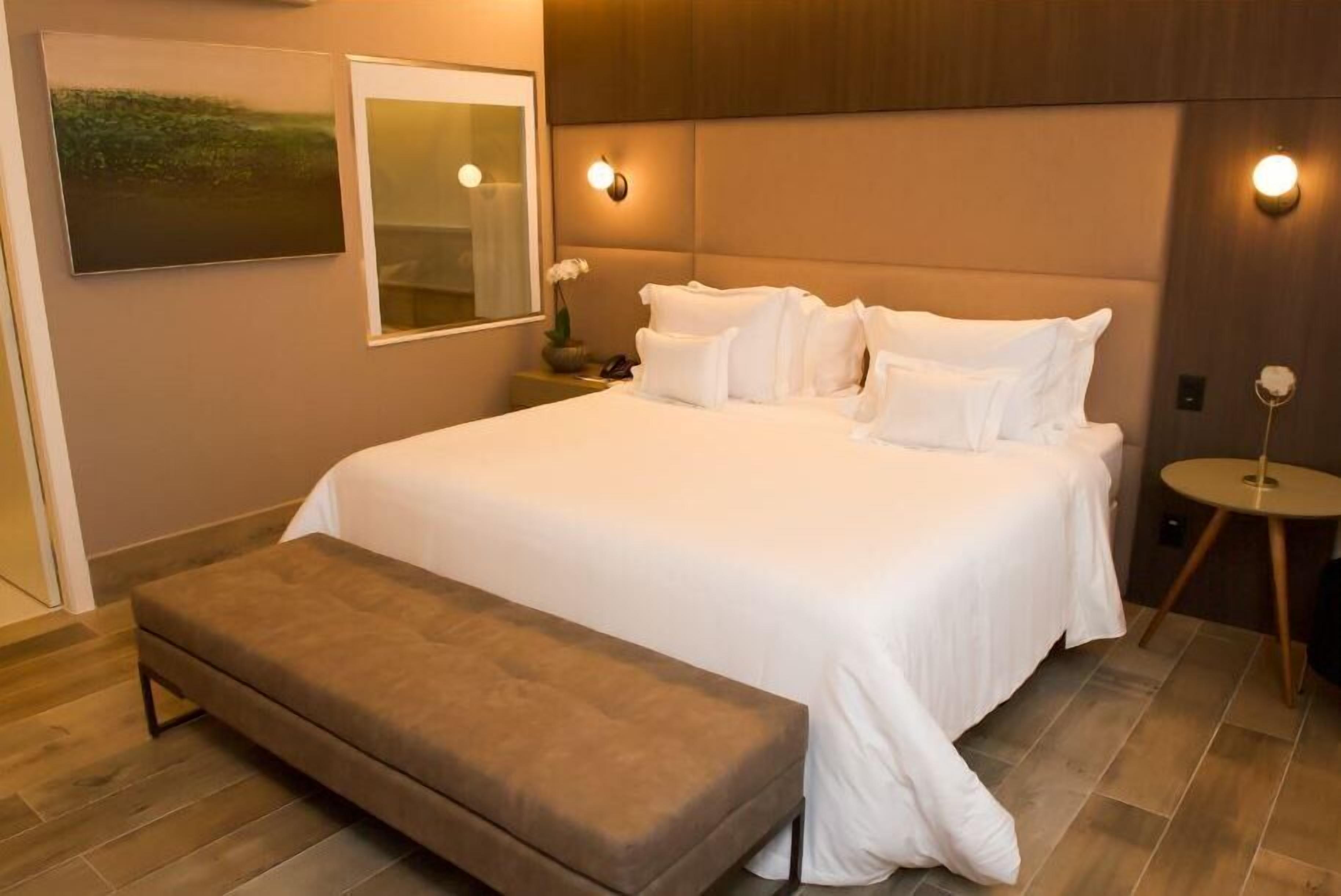 A neatly arranged bedroom with a large bed, a bench, a bedside table, and a wall-mounted light fixture.