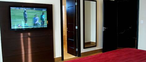 Una habitación de hotel con una televisión montada en la pared, un espejo grande y una colcha roja.