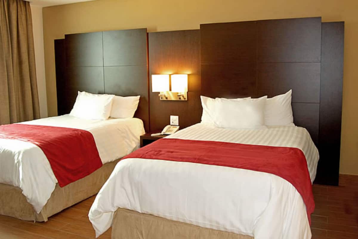 A hotel room with two beds, each with white bedding and red accents, a nightstand with a phone, and wall-mounted lighting.