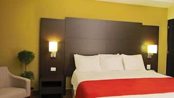 A hotel room with a bed, two bedside lamps, a small table, a chair, and a potted plant.