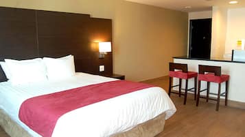 A hotel room with a large bed, a nightstand, a desk with two chairs, and a kitchenette.