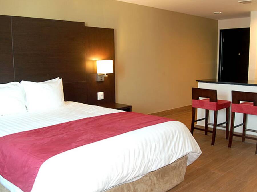 A hotel room with a large bed, a nightstand, a desk with two chairs, and a kitchenette.