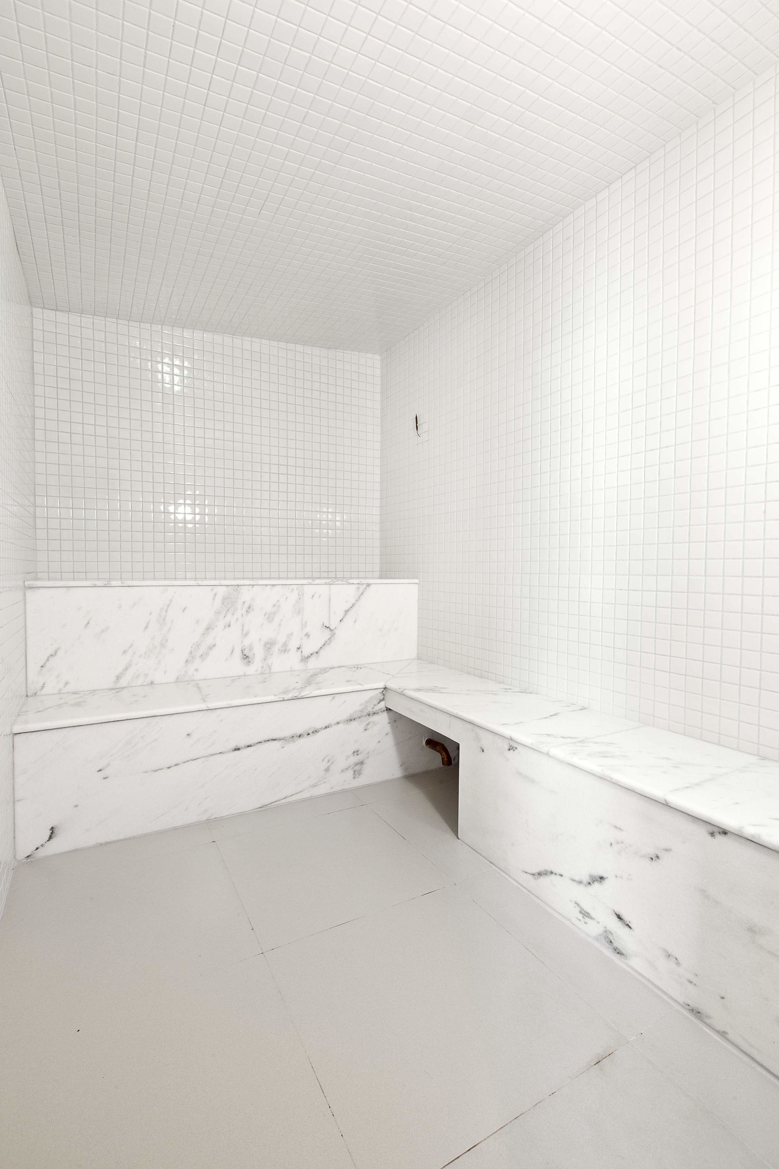 A bathroom with white tiled walls and floor, a marble bench, and a shower area.