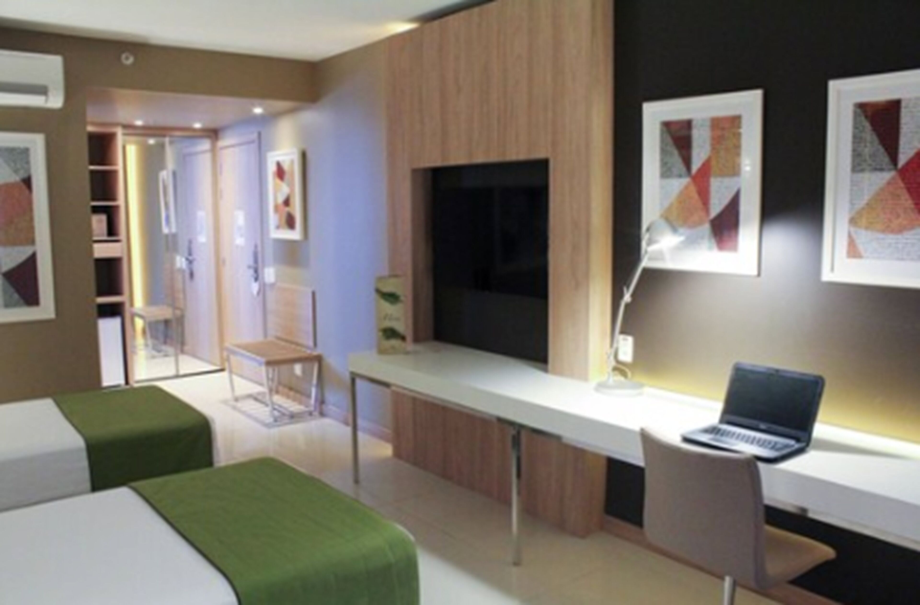 A modern hotel room with two beds, a desk with a laptop, a TV, and artwork on the walls.