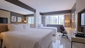 A hotel room with two beds, a desk, a chair, a TV, and large windows.
