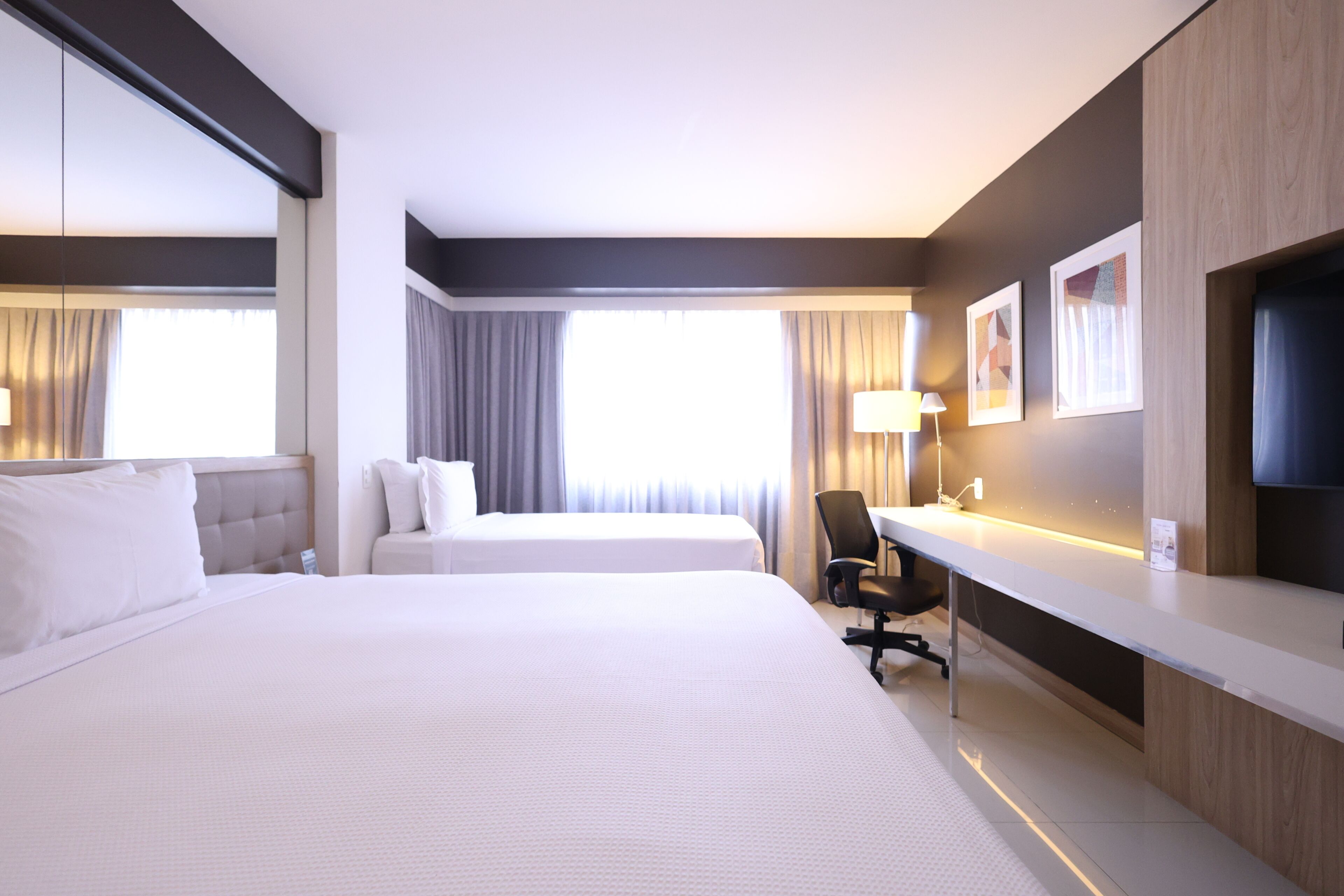 A hotel room with two beds, a desk, a chair, a TV, and artwork on the wall.