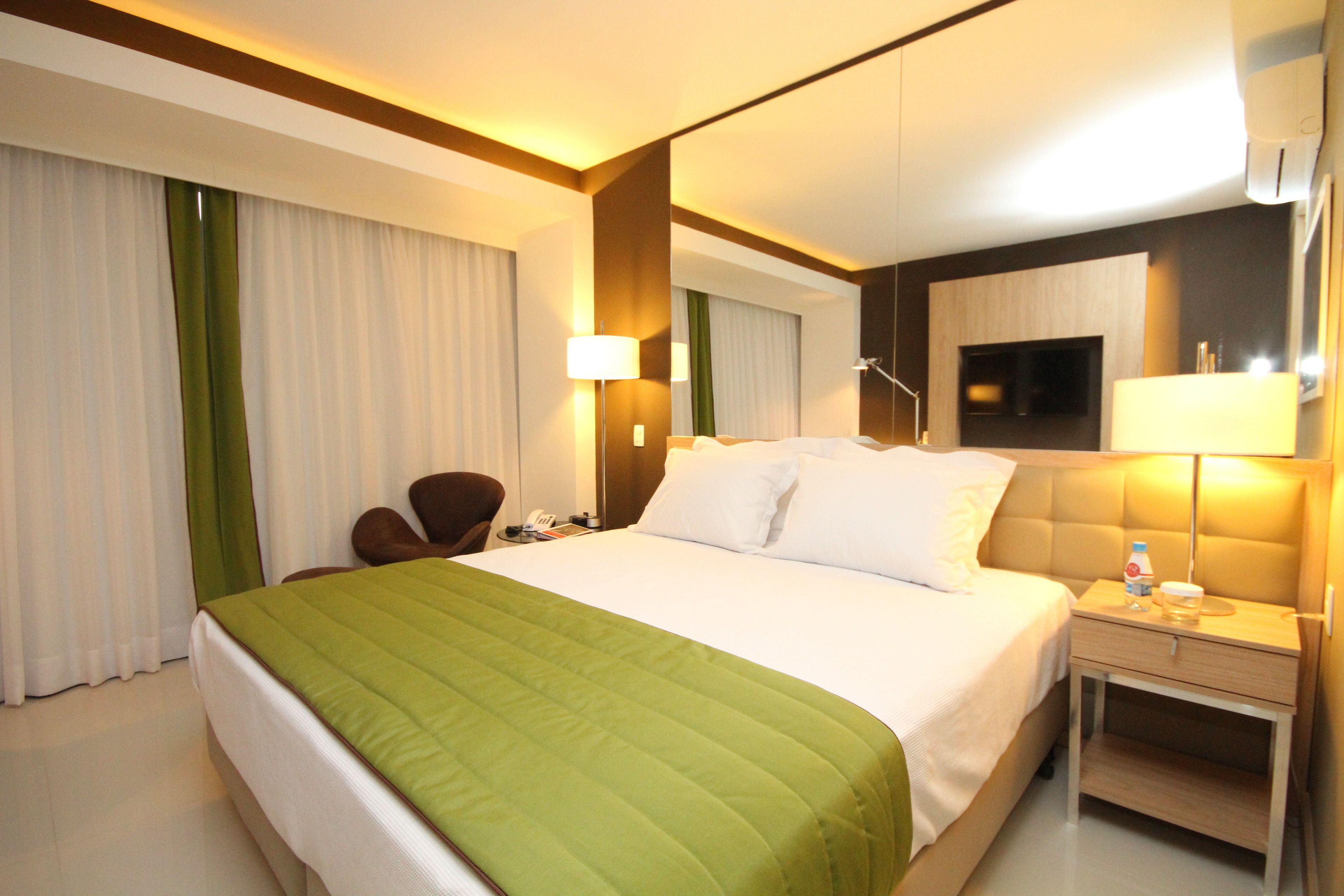 Habitación de hotel con una cama grande, una mesita de noche con lámpara, una silla, un escritorio y un televisor en la pared.