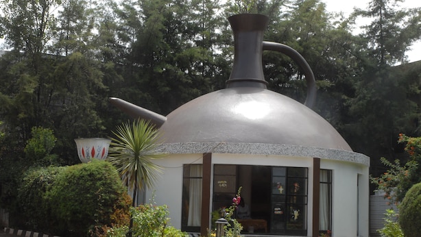 Een gebouw met een grote, koepelvormige structuur die op een theepot lijkt, omgeven door weelderige begroeiing en een verhard entreegebied.