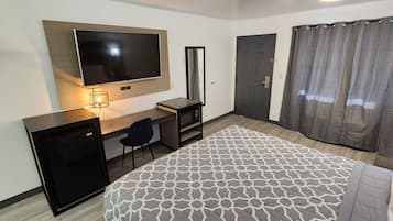 A hotel room with a bed, a desk with a computer, a TV mounted on the wall, a door, and a shower curtain.