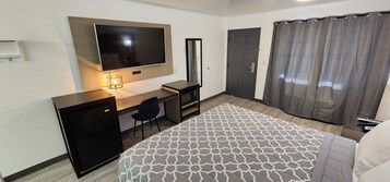 Habitación de hotel con cama, escritorio con computadora, televisor en la pared, puerta y cortina de ducha.