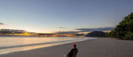 A person sitting on a chair on a sandy beach with a view of the ocean and a distant mountain range.