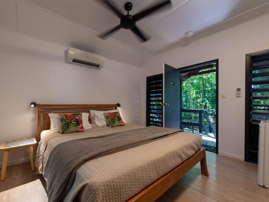 Un dormitorio con cama, ventilador de techo, equipo de aire acondicionado, un taburete pequeño y ventana con vista a la vegetación.