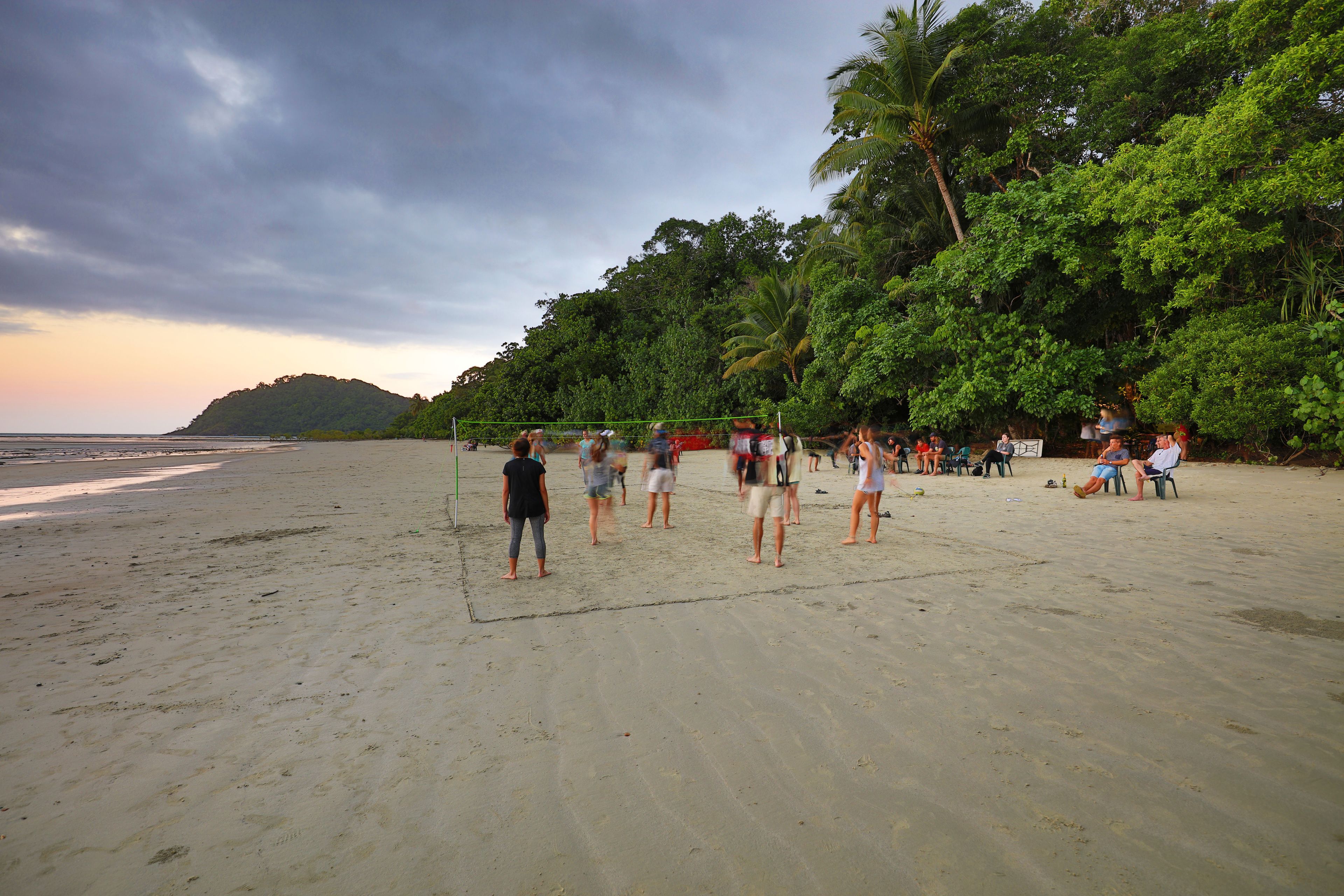 A beach volleyball court with players and spectators, surrounded by palm trees and a view of the ocean.