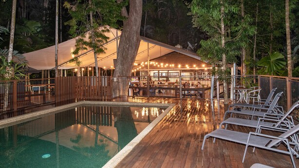 Zona de piscina con sillones reclinables, terraza de madera y área de descanso techada con luces colgantes.