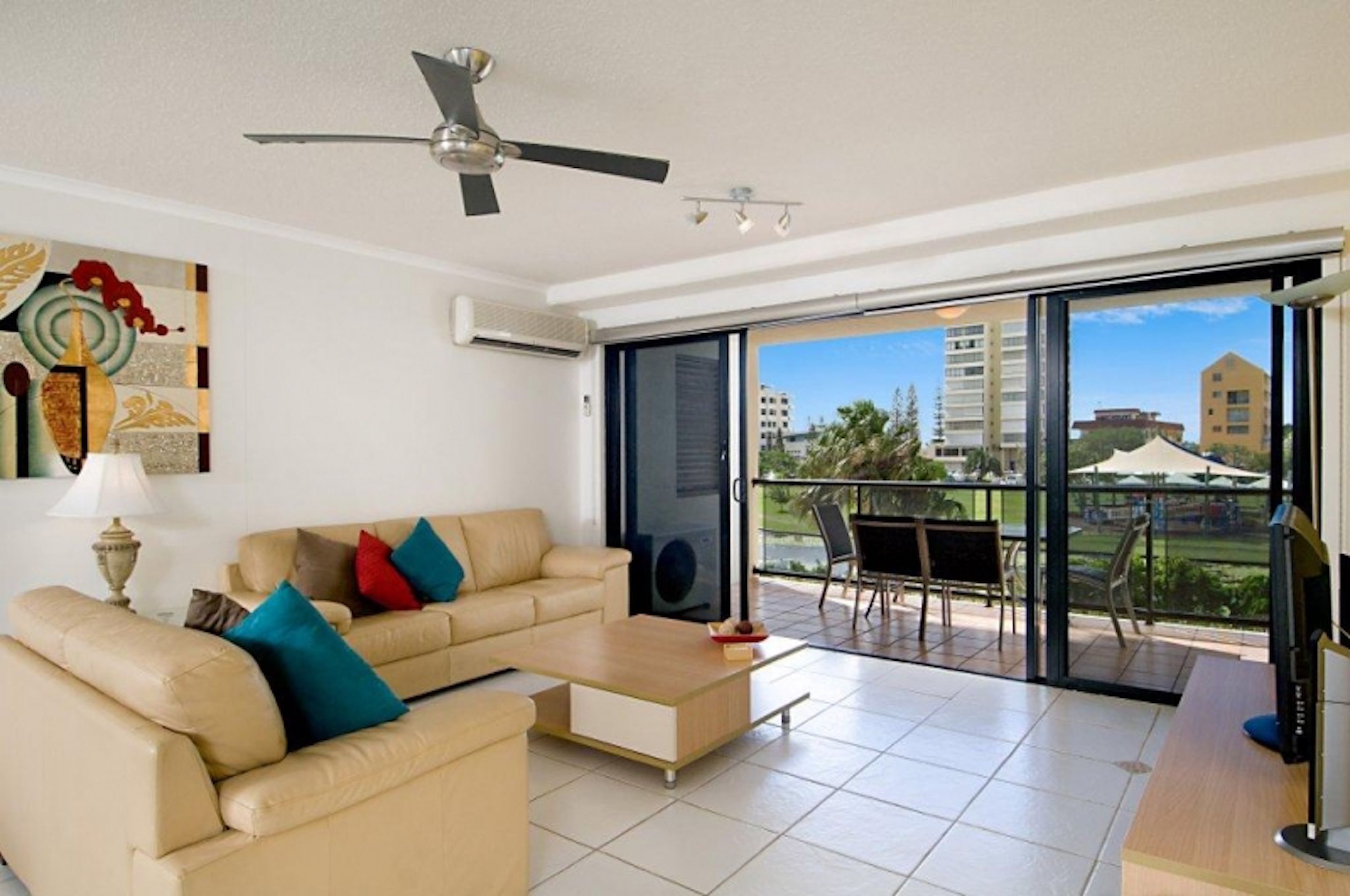A modern living room with a sofa, coffee table, and a ceiling fan. There is a sliding glass door leading to an outdoor area with a table and chairs.
