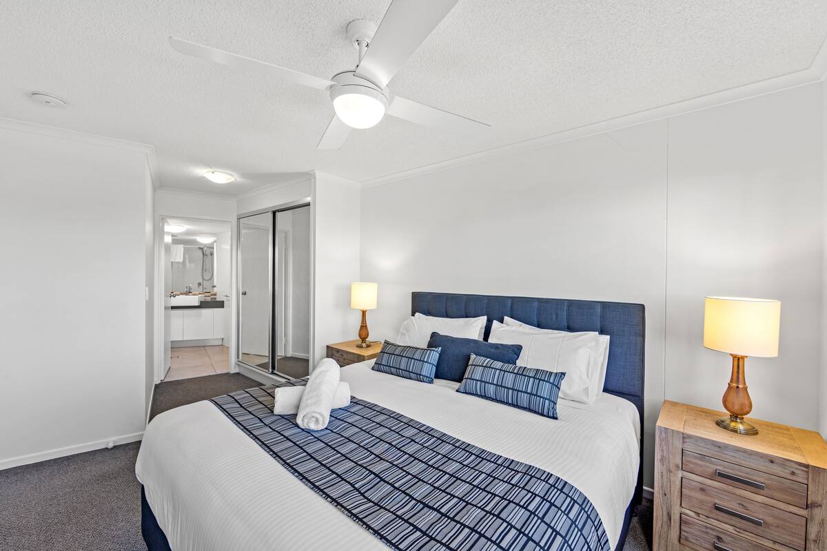 A hotel room with a large bed, a ceiling fan, a bedside table with a lamp, and a bathroom visible through an open door.
