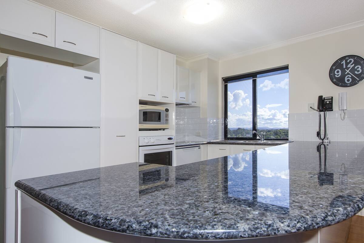 A kitchen with white cabinets, a granite countertop, a microwave, an oven, and a window with a view of the sky.